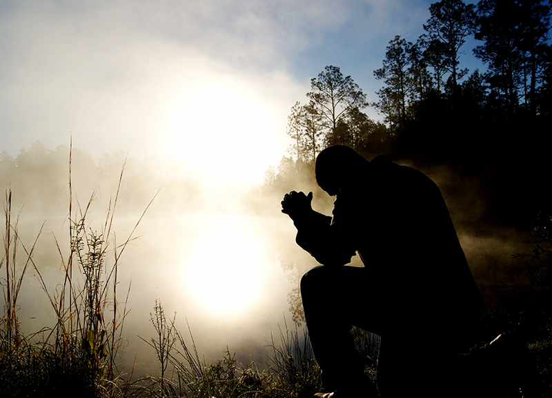 Mies istuu auringonnousussa kasteisella niityllä ja rukoilee.